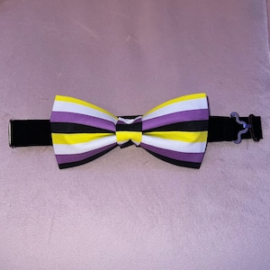 May include: A black bow tie with purple, yellow, and white stripes. The bow tie is attached to a black elastic band.