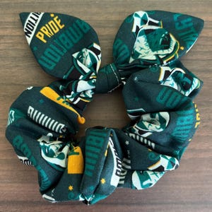 May include: A dark green fabric hair scrunchie featuring a repeating pattern of yellow and white text that includes the words PRIDE and AMBITION, along with small star graphics and stylized illustrations of a badger mascot on the cotton material.