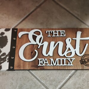 K&ouml;nnte beinhalten: Ein Schild aus Holz mit einem Kuhfellrand. Das Schild tr&auml;gt die Aufschrift "The Ernst Family Est. 2000" in wei&szlig;en Buchstaben.