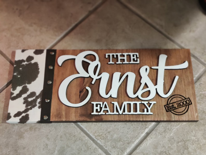 K&ouml;nnte beinhalten: Ein Schild aus Holz mit einem Kuhfellrand. Das Schild tr&auml;gt die Aufschrift "The Ernst Family Est. 2000" in wei&szlig;en Buchstaben.