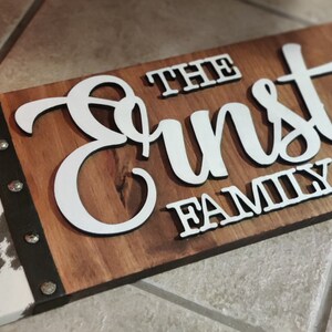 K&ouml;nnte beinhalten: Ein Holzschild mit einem Kuhfellmuster auf der linken Seite. Das Schild tr&auml;gt die Aufschrift "The Ernst Family Est. 2000" in wei&szlig;en Buchstaben.
