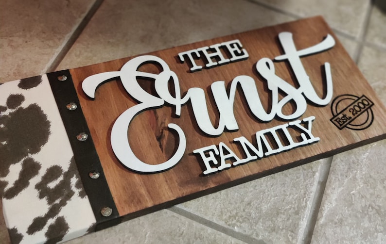 K&ouml;nnte beinhalten: Ein Holzschild mit einem Kuhfellmuster auf der linken Seite. Das Schild tr&auml;gt die Aufschrift "The Ernst Family Est. 2000" in wei&szlig;en Buchstaben.