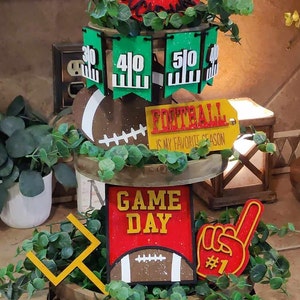 May include: A three-tiered wooden stand decorated with football-themed decorations. The top tier features a red circle with the words "Tailgater Tackles" in black lettering. The middle tier has green and white pennants with numbers 30, 40, and 50, a brown football, and a yellow sign that reads "Football is my favorite season". The bottom tier has a red sign with the words "Game Day" in yellow lettering, a brown football, a yellow goal post, and a red hand with the number 1.