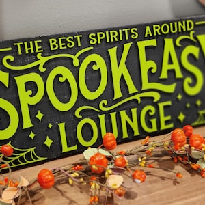May include: A black wooden sign with a green spiderweb design and the text "The Best Spirits Around Spook Easy Lounge".