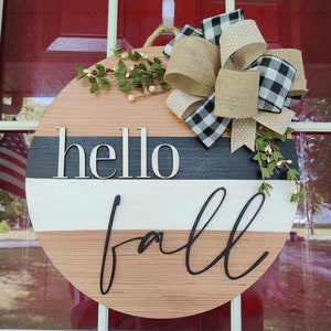 May include: A round wooden door hanger with a black and white striped design. The words "hello fall" are written in black script on the hanger. A brown and black checkered bow with white polka dots is attached to the top of the hanger.
