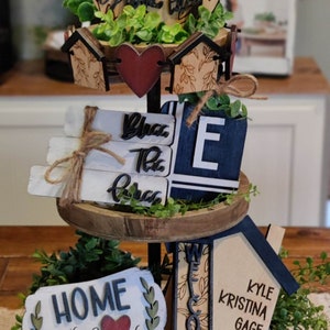 May include: A three-tiered wooden tiered tray with a farmhouse theme. The top tier features a round wooden sign with the words "Love" and a red heart. The middle tier has a sign that says "Bless the Mess" and a blue sign with the letter "E". The bottom tier has a sign that says "Home is where the heart is" and a sign that says "Welcome". The tray is decorated with greenery.