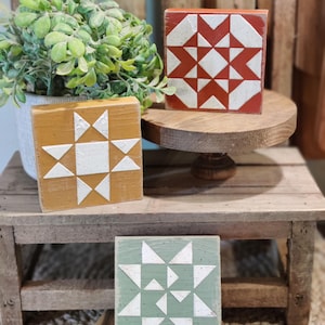 May include: Three square wooden blocks featuring painted geometric star patterns in burnt orange, mustard yellow, and sage green. Each block displays a white, speckled star design on its front face, showcasing a distinct arrangement of triangles.