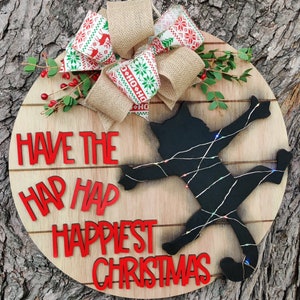 May include: A round wooden sign with a black silhouette of a cat tangled in Christmas lights. The sign says "Have the hap happiest Christmas". The sign is decorated with a red and green plaid bow and a burlap bow.