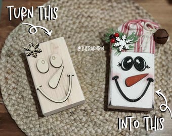 May include: Wooden snowman craft kit with a natural wood base and separate face pieces. The finished snowman has a painted white face, a carrot nose, and a striped hat. The text "TURN THIS INTO THIS" is also in the image.