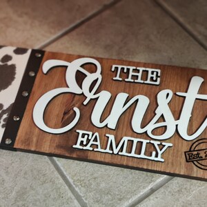 K&ouml;nnte beinhalten: Ein Holzschild mit einem Kuhfellmuster auf der linken Seite. Das Schild tr&auml;gt die Aufschrift "The Ernst Family Est. 2000" in wei&szlig;en Buchstaben.