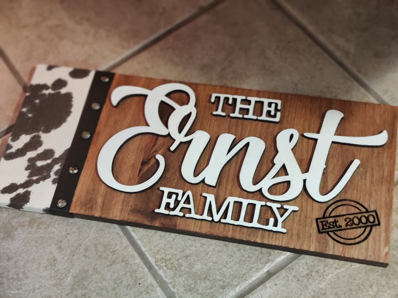 K&ouml;nnte beinhalten: Ein Holzschild mit einem Kuhfellmuster auf der linken Seite. Das Schild tr&auml;gt die Aufschrift "The Ernst Family Est. 2000" in wei&szlig;en Buchstaben.