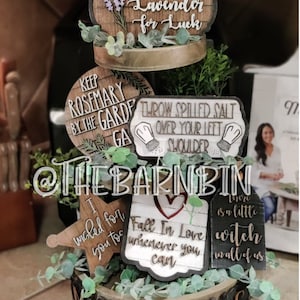 May include: A tiered wooden tray with various signs and decorations. The signs have sayings such as "Plant Lavender for Luck", "Keep Rosemary by the Garden Gate", "Throw Spilled Salt Over Your Left Shoulder", "I Wished for You Too", "Fall in Love Wherever You Can", and "There is a Little Witch in All of Us". The tray is decorated with greenery and has the word "MAGIC" spelled out on the bottom.