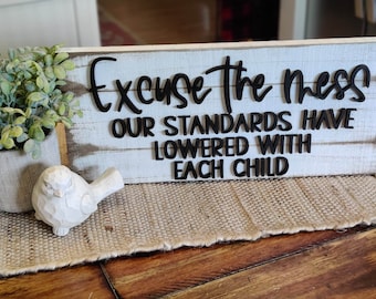 May include: White wooden sign with black lettering that reads "Excuse the mess our standards have lowered with each child".