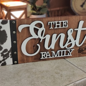 K&ouml;nnte beinhalten: Ein Holzschild mit einem Kuhfellhintergrund. Das Schild tr&auml;gt die Aufschrift "The Ernst Family Est. 2000" in wei&szlig;en Buchstaben.
