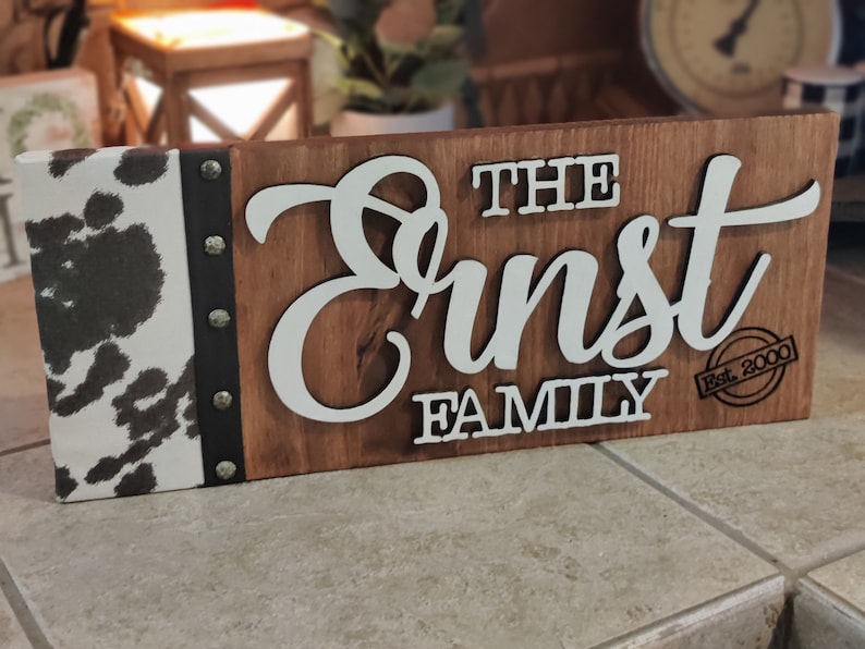 K&ouml;nnte beinhalten: Ein Holzschild mit einem Kuhfellhintergrund. Das Schild tr&auml;gt die Aufschrift "The Ernst Family Est. 2000" in wei&szlig;en Buchstaben.