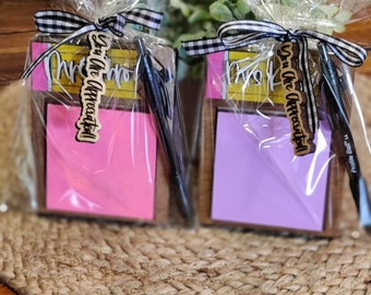 May include: Two gift sets, each containing a notepad, pen, and a wooden tag that reads "You Are Appreciated!" The notepads are pink and purple. The sets are wrapped in clear cellophane and tied with a black and white checkered ribbon.