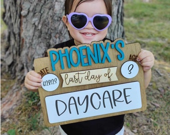 May include: A wooden sign with the text "PHOENIX'S last day of DAYCARE" and the date "6/29/23" and age "3" written on it. A child wearing purple heart-shaped sunglasses is holding the sign.
