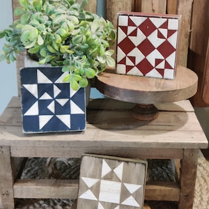 May include: Three square wooden blocks featuring geometric quilt-inspired star patterns in red and white, navy blue and white, and grey and white. The blocks are displayed on a small wooden riser alongside a potted green plant.