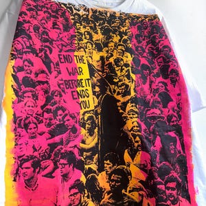 May include: White t-shirt with a vibrant graphic print featuring a crowd of people in shades of pink, yellow, and black. The text "END THE WAR BEFORE IT ENDS YOU" is prominently displayed. The shirt is hanging on a white hanger.