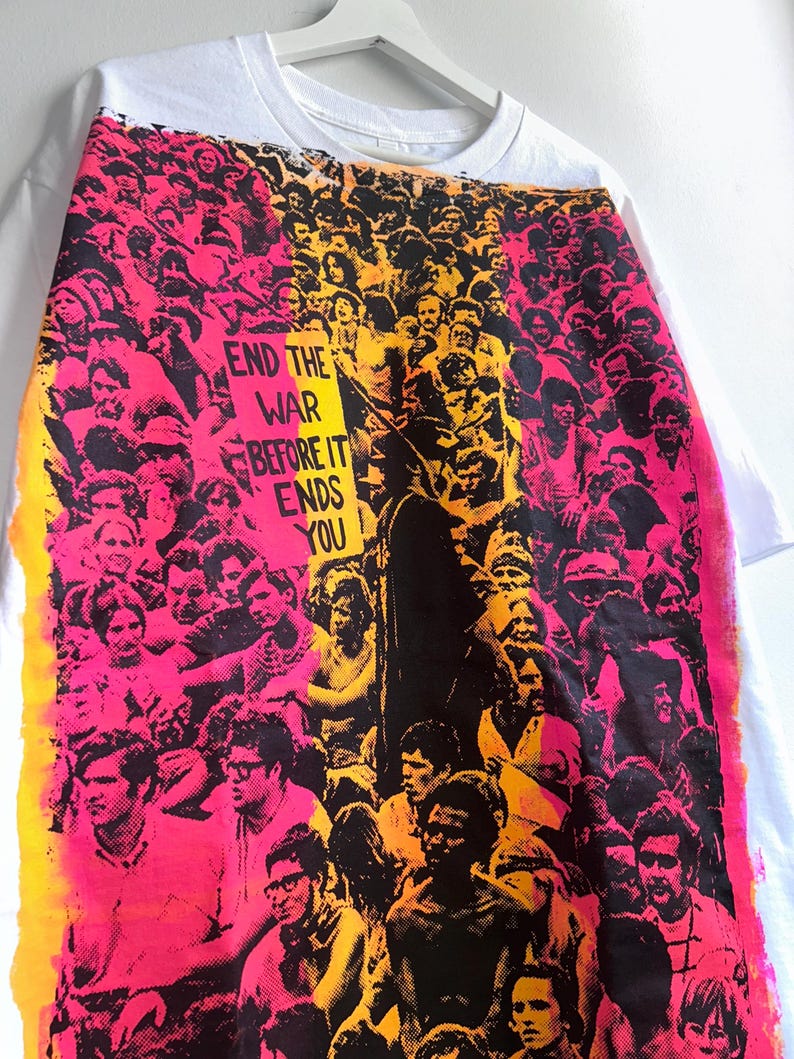 May include: White t-shirt with a vibrant graphic print featuring a crowd of people in shades of pink, yellow, and black. The text "END THE WAR BEFORE IT ENDS YOU" is prominently displayed. The shirt is hanging on a white hanger.