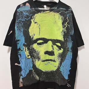 Puede incluir: Camiseta negra con un gran diseño gráfico en verde neón y negro del monstruo de Frankenstein. El diseño incluye rayos azules y una estética vintage y desgastada. La camiseta está colgada en una percha blanca.