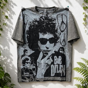 Può includere: Una t-shirt grigia con effetto lavaggio acido caratterizzata da una grande grafica in bianco e nero di Bob Dylan con occhiali da sole. Il design include simboli di pace, testo scritto a mano e le frasi Bob Dylan e Knockin' on Heaven's Door.