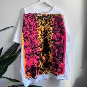 May include: White t-shirt featuring a vibrant graphic of a crowd in shades of pink, orange, and black. The graphic includes the text "END THE WAR BEFORE IT ENDS YOU" on a sign. The shirt is hanging on a white hanger.