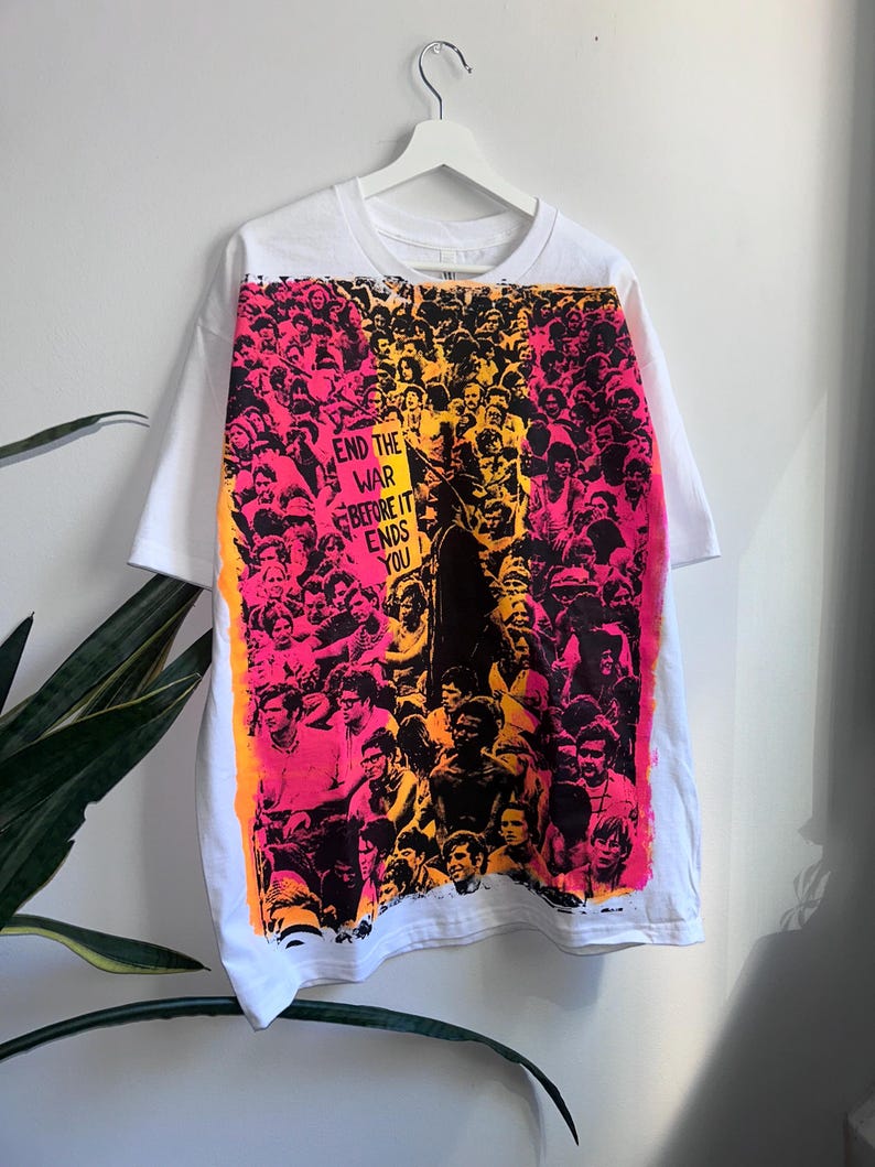 May include: White t-shirt featuring a vibrant graphic of a crowd in shades of pink, orange, and black. The graphic includes the text "END THE WAR BEFORE IT ENDS YOU" on a sign. The shirt is hanging on a white hanger.