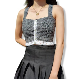 May include: A gray and white crochet tank top with a button-up front and a white crocheted trim. The top is worn with a black pleated skirt.