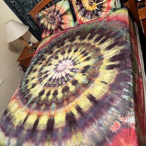May include: A bed with a tie-dye patterned duvet cover and two matching pillowcases. The duvet cover features a large, circular design in shades of yellow, red, purple, and black. The pillowcases have a smaller, spiral design in similar colors.