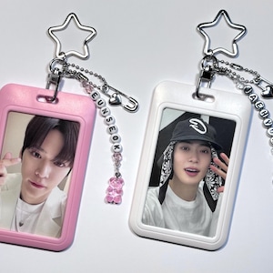 Pode incluir: Dois porta-cartões fotográficos retangulares de plástico, um rosa e um branco, cada um com um porta-chaves de metal em forma de estrela. Incluem correntes de contas com letras que formam EUNSEOK e JAEHYUN, com pequenos pingentes de ursinho.