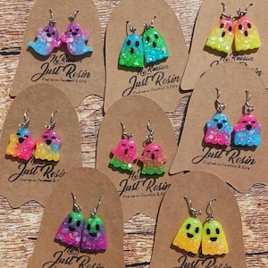 May include: Eight pairs of ghost-shaped resin earrings in various bright, glittery color gradients. Each pair features a unique color combination, such as pink and blue, green and yellow, or orange and yellow, all attached to silver-toned metal hooks.