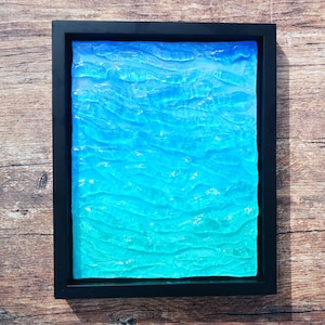 May include: A framed artwork featuring a textured, abstract design in shades of blue and turquoise, resembling water. The artwork is encased in a black rectangular frame, creating a striking contrast against the wooden background.