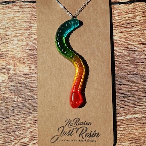 May include: A colorful gummy worm pendant necklace. The pendant is a gradient of colors, transitioning from red to orange, yellow, green, and blue. The necklace is on a silver chain and displayed on a brown card with the text "No Reason Just Resin".