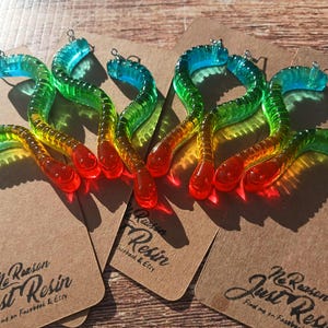K&ouml;nnte beinhalten: Bunte, wurmf&ouml;rmige Charms aus Kunstharz in Rot, Gelb, Gr&uuml;n und Blau. Jeder Charm hat eine kleine Metallschlaufe. Die Charms werden auf braunen Karten mit dem Text "No Reason Just Resin" pr&auml;sentiert.