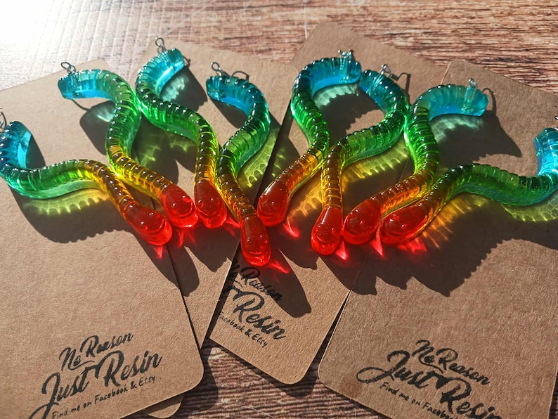K&ouml;nnte beinhalten: Bunte, wurmf&ouml;rmige Charms aus Kunstharz in Rot, Gelb, Gr&uuml;n und Blau. Jeder Charm hat eine kleine Metallschlaufe. Die Charms werden auf braunen Karten mit dem Text "No Reason Just Resin" pr&auml;sentiert.