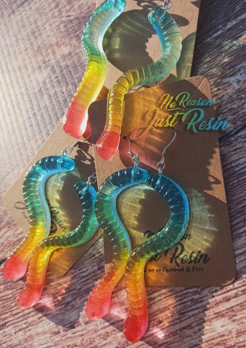 K&ouml;nnte beinhalten: Bunte Wurm-Ohrringe aus Kunstharz mit einem Farbverlauf von Blau, Gr&uuml;n, Gelb und Rot. Die Ohrringe haben silberne Haken und werden auf einer braunen Karte mit dem Text "No Reason Just Resin" pr&auml;sentiert.