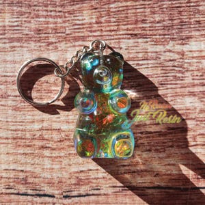 May include: A colorful, iridescent gummy bear keychain. The bear is made of resin and has a silver keyring and chain. The bear's colors include blue, green, orange, and yellow. The background is a wooden surface.