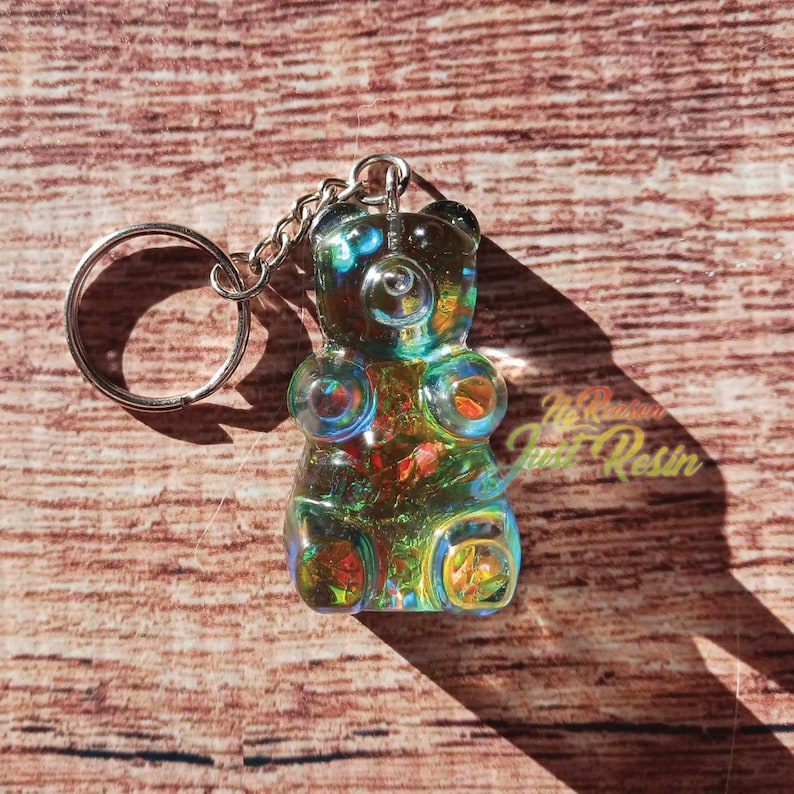 May include: A colorful, iridescent gummy bear keychain. The bear is made of resin and has a silver keyring and chain. The bear's colors include blue, green, orange, and yellow. The background is a wooden surface.