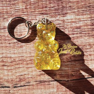 May include: A yellow gummy bear keychain with glitter and a silver keyring. The keychain has the text "No Reason Just Resin" on the bottom right corner. The background is a wooden surface.