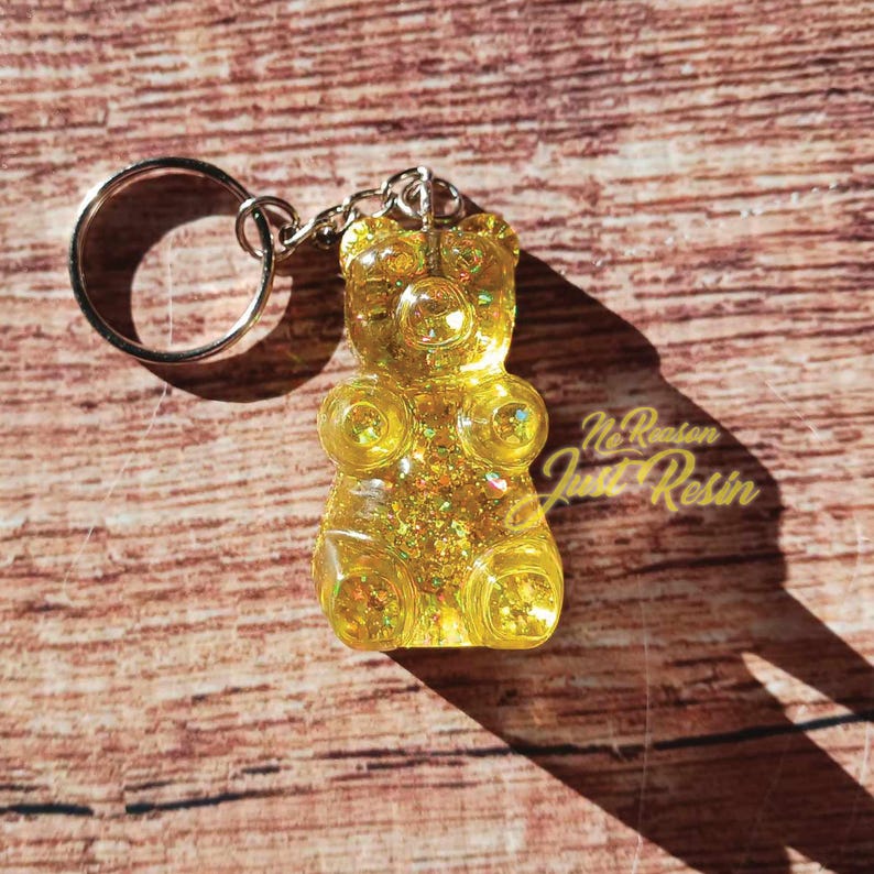 May include: A yellow gummy bear keychain with glitter and a silver keyring. The keychain has the text "No Reason Just Resin" on the bottom right corner. The background is a wooden surface.