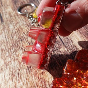 May include: A clear gummy bear keychain with red glitter accents. The gummy bear is suspended from a silver keyring. The image is a close-up shot, highlighting the details of the resin and glitter.