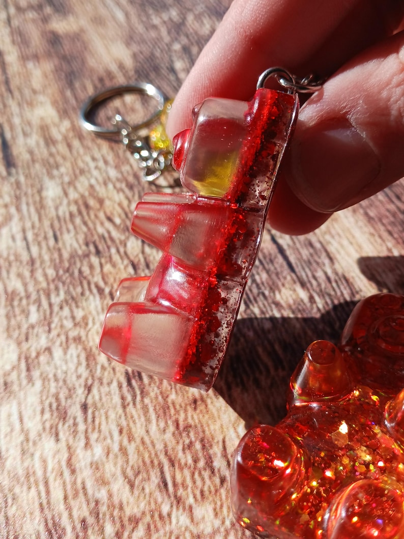 May include: A clear gummy bear keychain with red glitter accents. The gummy bear is suspended from a silver keyring. The image is a close-up shot, highlighting the details of the resin and glitter.