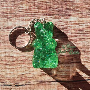 May include: A green gummy bear keychain made of resin. The keychain has a silver keyring and chain. The gummy bear is filled with glitter and has the text "No Reason Just Resin" in the bottom right corner.