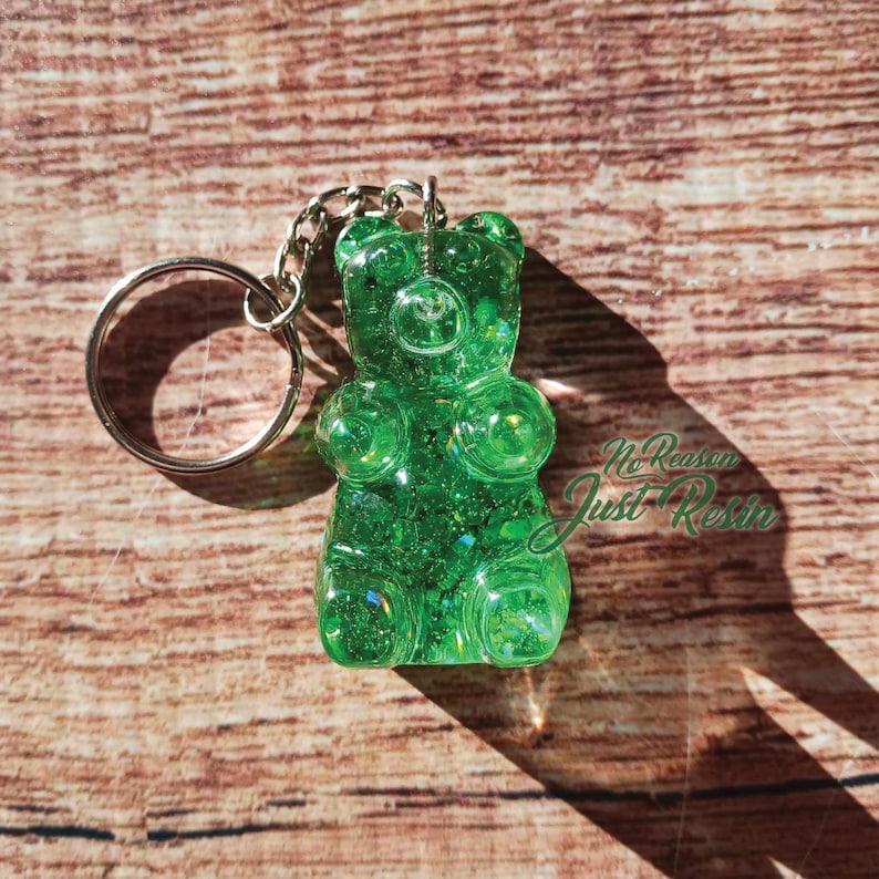 May include: A green gummy bear keychain made of resin. The keychain has a silver keyring and chain. The gummy bear is filled with glitter and has the text "No Reason Just Resin" in the bottom right corner.