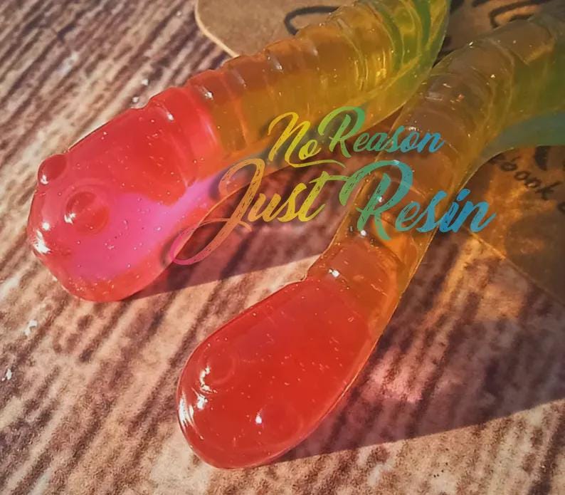 K&ouml;nnte beinhalten: Zwei Kunstharzw&uuml;rmer mit einem Farbverlauf aus Rot, Rosa, Gelb und Gr&uuml;n. Die W&uuml;rmer haben eine gl&auml;nzende Oberfl&auml;che und liegen auf einer strukturierten Oberfl&auml;che. Der Text "No Reason Just Resin" ist auf den W&uuml;rmern zu sehen.