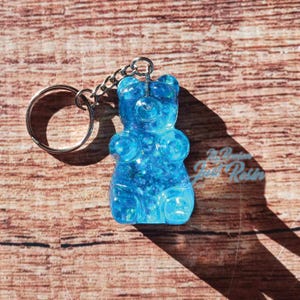 May include: A blue gummy bear keychain made of resin. The keychain has a silver chain and a silver key ring. The gummy bear is filled with glitter and has a glossy finish. The keychain is on a wooden surface.