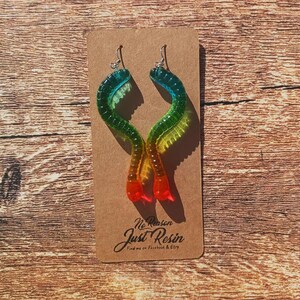 K&ouml;nnte beinhalten: Ein Paar bunte Harzohrringe in wurmartigem Design. Die Ohrringe wechseln von Blau zu Gr&uuml;n, Gelb und Rot. Die Ohrringe werden auf einer braunen Karte mit dem Text "No Reason Just Resin" pr&auml;sentiert.