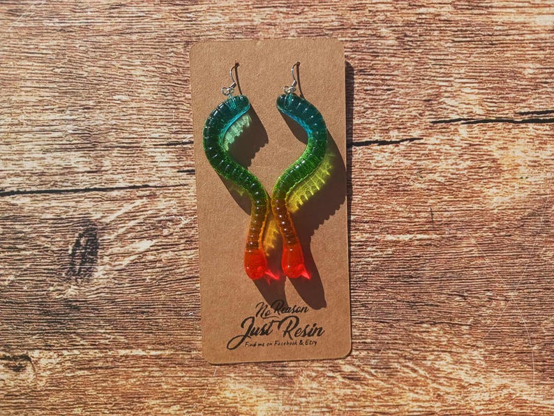 K&ouml;nnte beinhalten: Ein Paar bunte Harzohrringe in wurmartigem Design. Die Ohrringe wechseln von Blau zu Gr&uuml;n, Gelb und Rot. Die Ohrringe werden auf einer braunen Karte mit dem Text "No Reason Just Resin" pr&auml;sentiert.
