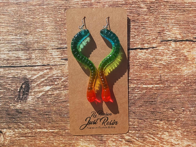 K&ouml;nnte beinhalten: Bunte Harz-Ohrringe in wurmartigem Design. Die Ohrringe wechseln von Blau zu Gr&uuml;n, Gelb, Orange und Rot. Die Ohrringe werden auf einer braunen Karte mit dem Text "No Reason Just Resin" pr&auml;sentiert.